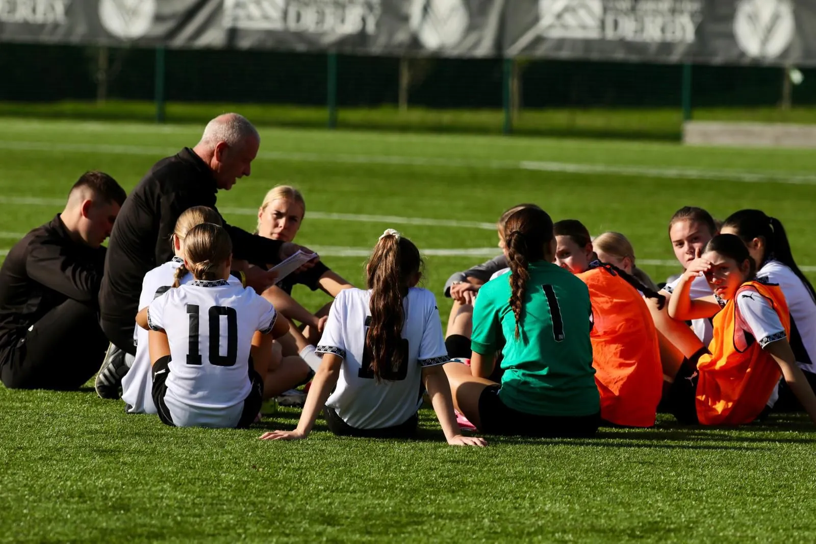 A coach crouching to talk with a group of players sitting on the pitch during a training session.
