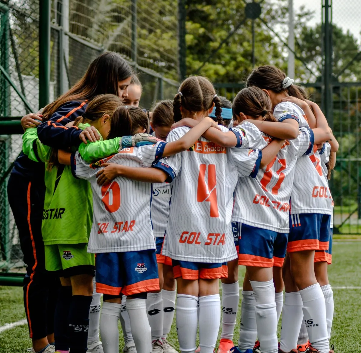 A girls' team in white and orange kit standing in a huddle on a pitch, arms around each other.